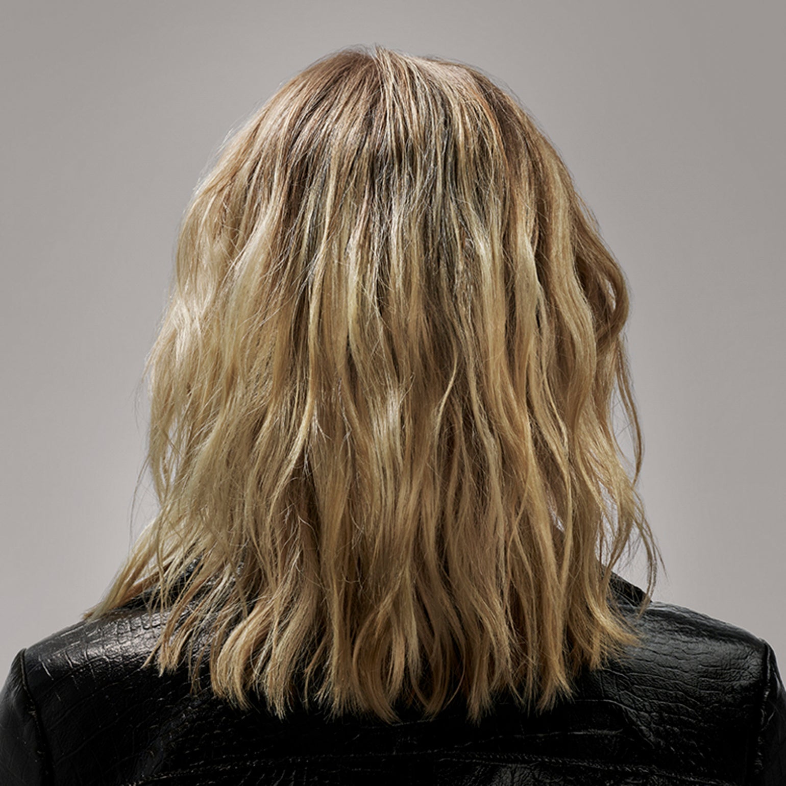 Back view of a person with long, wavy blonde hair wearing a black leather jacket on a gray background