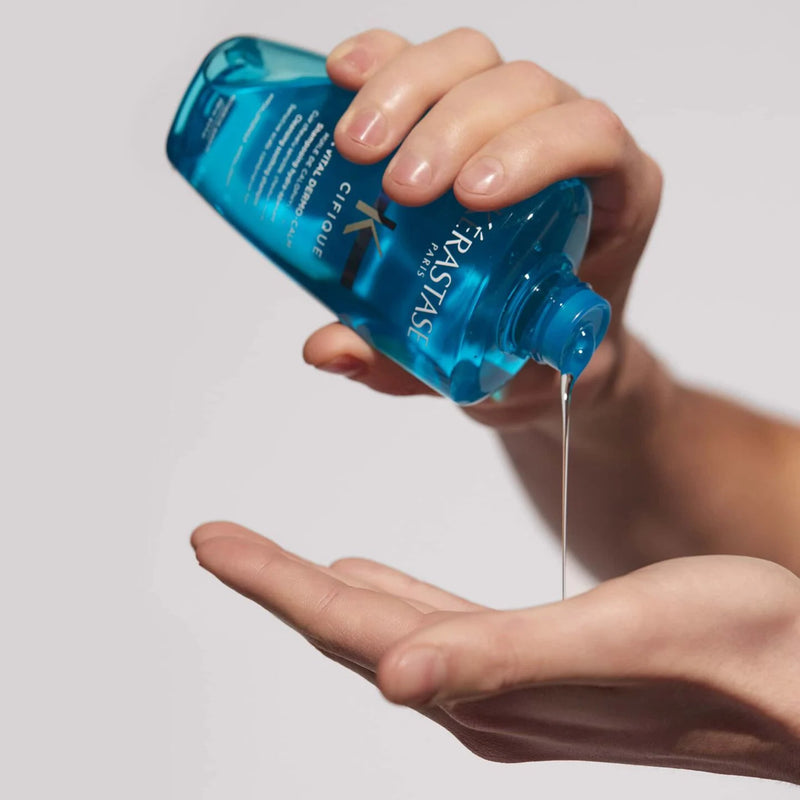 Hand holding a blue bottle of Dermo Cal oil over another hand with oil dripping.
