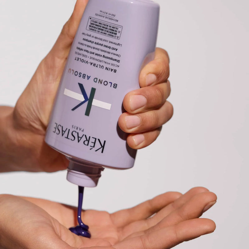 Hand holding a bottle of Kerastase product with purple liquid being dispensed onto the palm.