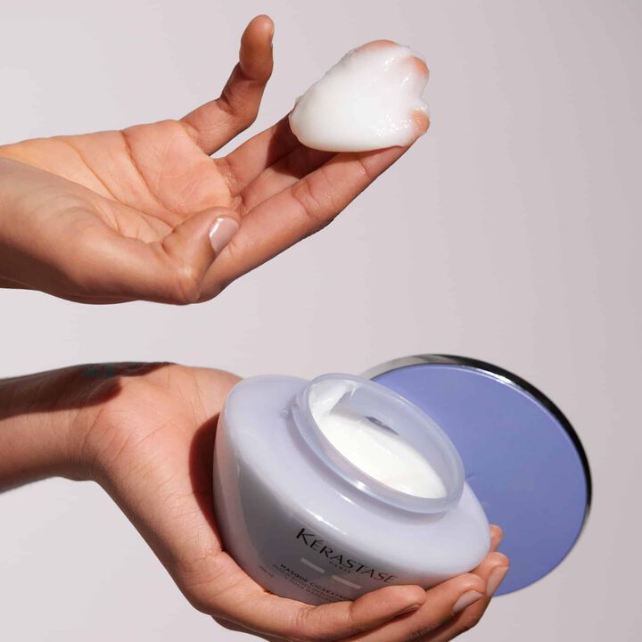 Person holding a jar of Kerastase product with part of the product gel texture in other hand on a plain background