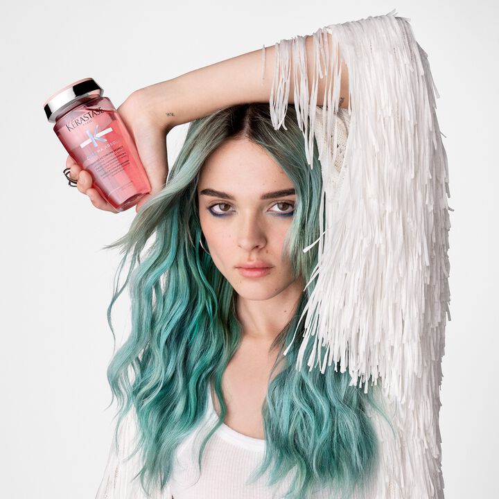 Person with teal hair holding a pink bottle against a white background
