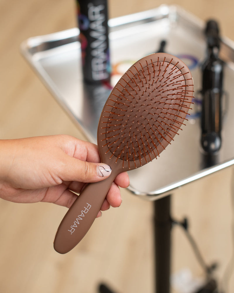 Brown hairbrush with 'FRAMAR' branding held by a hand, with a blurred background of a magazine and hair products.