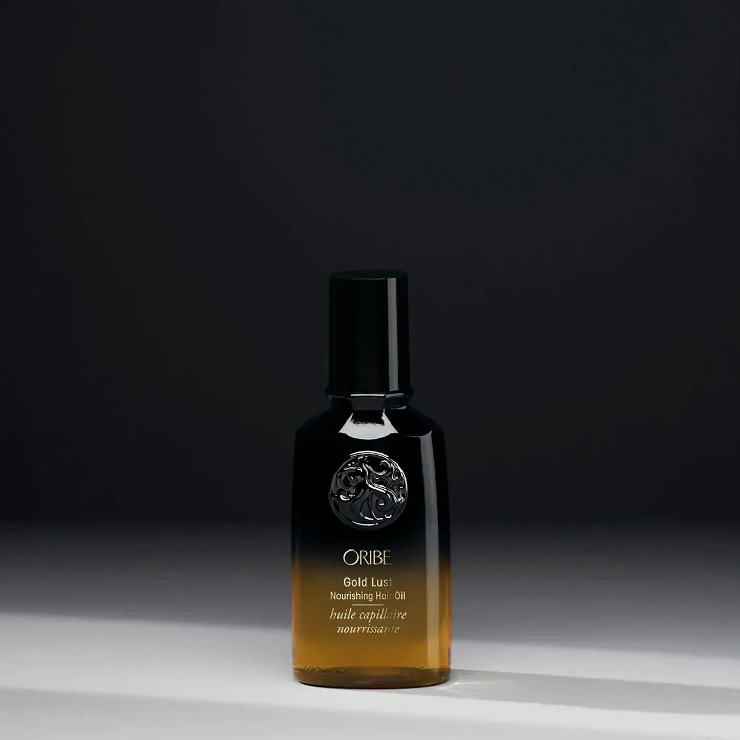 A bottle of ORIBE Gold Lust Nourishing Hair Oil with a dark cap and a clear, amber-coloured liquid visible through the bottle, set against a dark background.