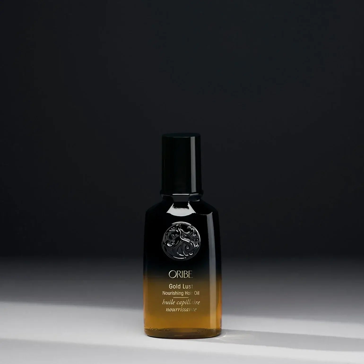 A bottle of ORIBE Gold Lust Nourishing Hair Oil with a dark cap and a clear, amber-coloured liquid visible through the bottle, set against a dark background.