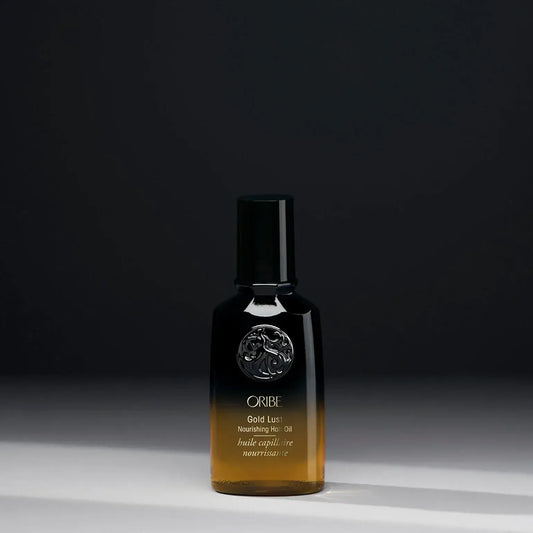 A bottle of ORIBE Gold Lust Nourishing Hair Oil with a dark cap and a clear, amber-coloured liquid visible through the bottle, set against a dark background.