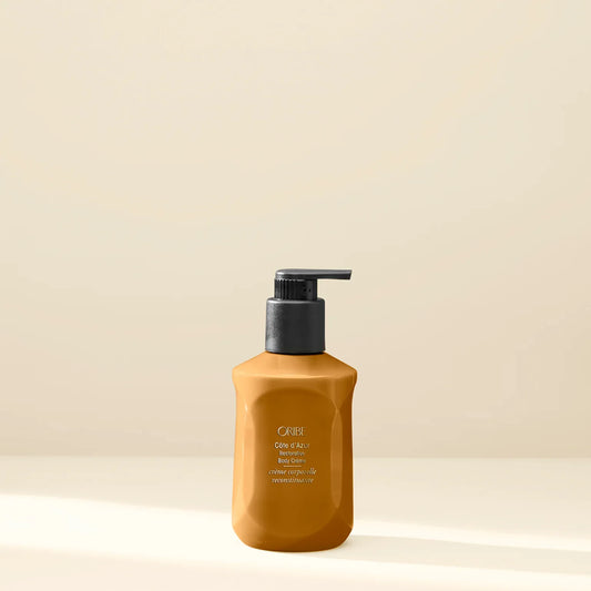 A pump bottle of ORIBE Côte d'Azur Restorative Body Crème with a rich orange color, displayed against a neutral background.