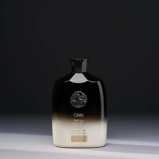 A bottle of ORIBE Gold Lust Repair & Restore Shampoo with a black to transparent gradient and gold details.