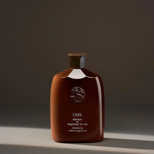 A bottle of ORIBE Shampoo for Magnificent Volume, with a dark brown label and cap, displayed against a shadowed background.